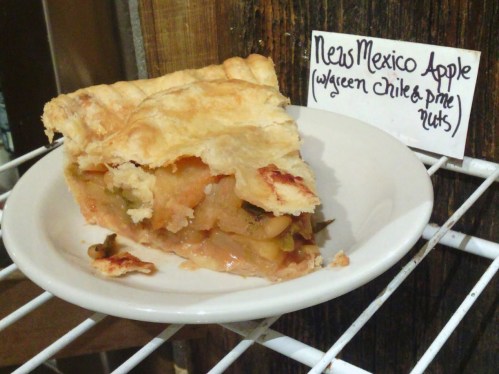 A slice of New Mexico apple pie with green chile and pine nuts on a white plate, displayed on a wire rack.