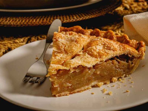 A slice of apple pie on a white plate, with a fork beside it, set on a textured tablecloth.