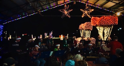 A festive outdoor scene with a band wearing Santa hats, surrounded by colorful holiday lights and decorations.