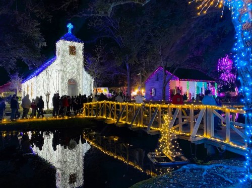 A festive scene at night featuring a lit church, colorful lights, and a bridge over a reflective pond with people gathered.