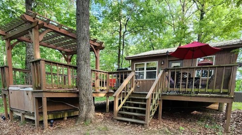 A wooden cabin surrounded by trees, featuring a deck with an umbrella and a hot tub on a separate platform.