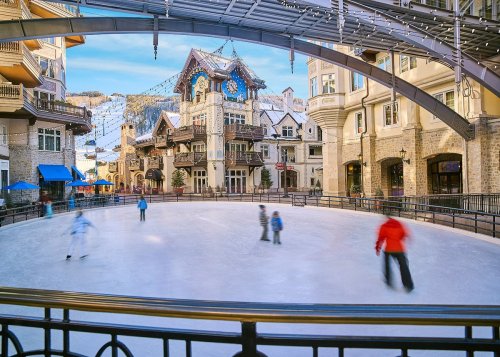 A scenic ice skating rink surrounded by charming buildings and mountains, with skaters gliding across the ice.