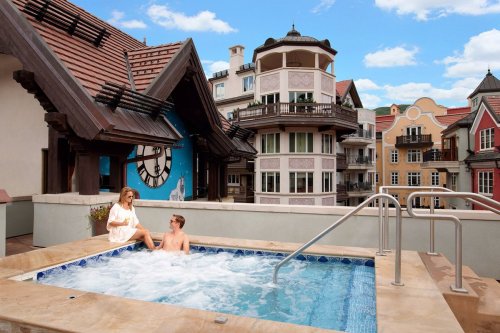 A couple relaxes in a rooftop hot tub with scenic buildings and a blue sky in the background.