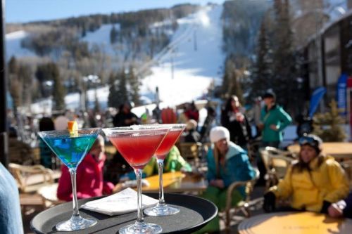Two colorful cocktails on a tray in the foreground, with a snowy mountain and skiers in the background.