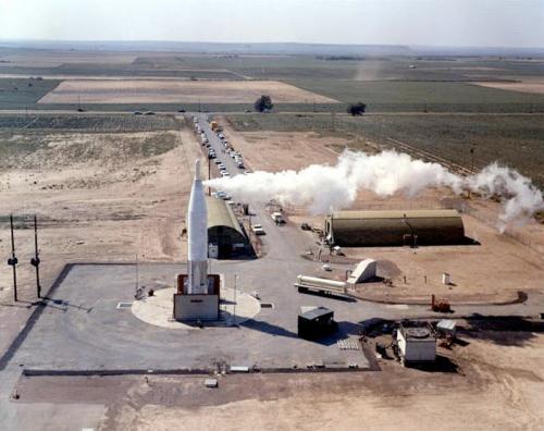 Aerial view of a missile launch site with smoke billowing from a rocket, surrounded by open fields and structures.