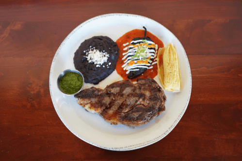 A plate featuring grilled chicken, black beans, a stuffed pepper, salsa, and a side of tamale with green sauce.