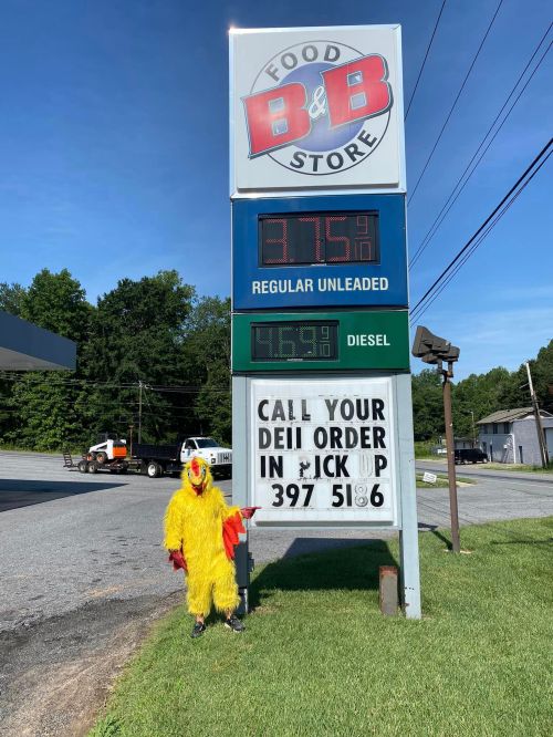 A person in a chicken costume stands in front of a gas station sign displaying fuel prices and a deli order number.