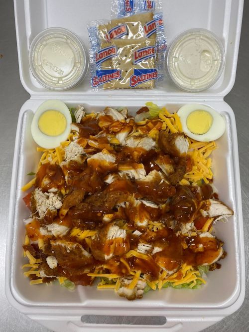 A takeout container filled with salad topped with chicken, cheese, barbecue sauce, boiled eggs, and crackers.