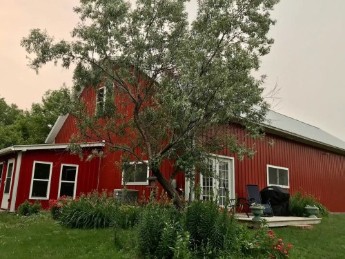 A red barn-style house surrounded by greenery and a large tree, with a patio and chairs visible in the foreground.