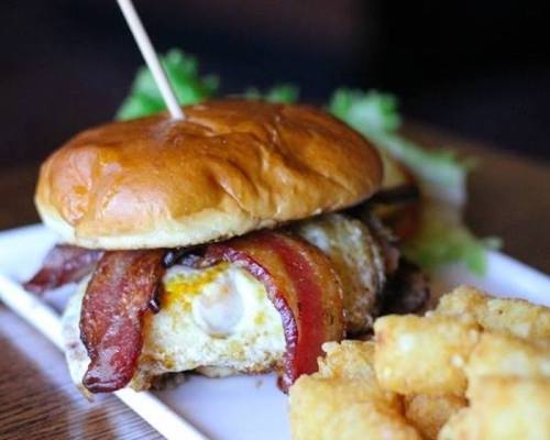 A juicy burger with bacon and a fried egg on a bun, served with crispy tater tots on the side.