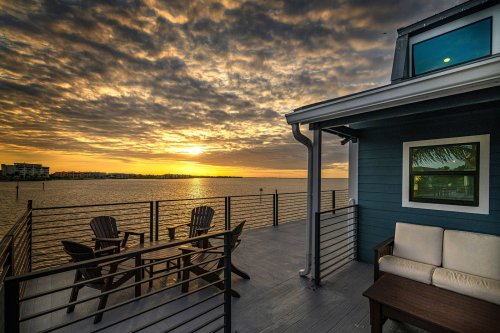 A serene waterfront view at sunset, featuring a cozy deck with chairs and a sofa, framed by dramatic clouds.