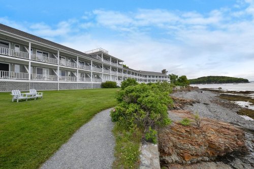 A coastal hotel with a grassy area and rocky shoreline, under a blue sky with scattered clouds.