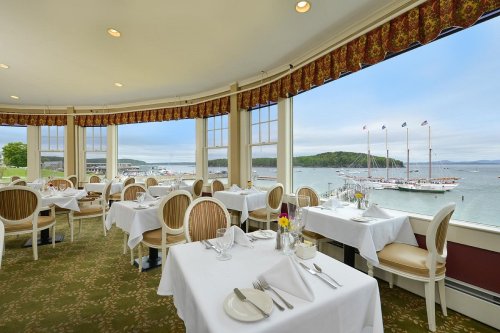 Elegant restaurant interior with white tablecloths, overlooking a scenic waterfront with boats and hills in the distance.