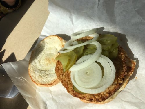 A fried chicken sandwich on a paper wrapper, topped with sliced onions and pickles, with the bottom bun beside it.