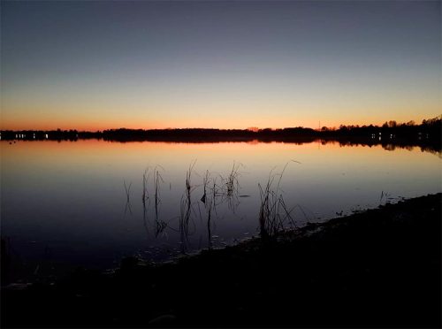 A serene lake at sunset, reflecting vibrant colors in the sky with silhouettes of grass along the shore.