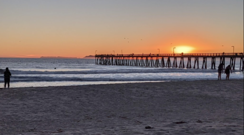 Sunset over a pier with silhouetted figures on the beach and gentle waves lapping at the shore.