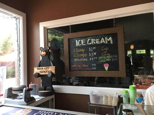 A chalkboard menu displays ice cream prices and options at a shop, with a bear statue nearby.