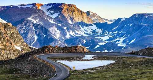 A winding road curves through a mountainous landscape with snow-capped peaks and green valleys under a clear sky.