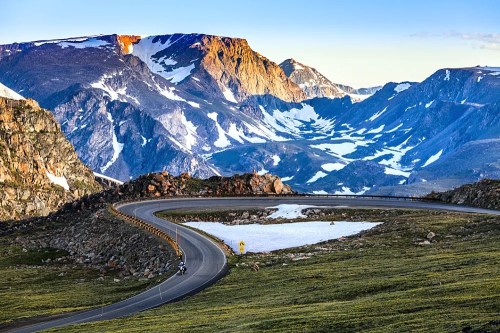A winding road curves through a mountainous landscape with snow-capped peaks and a clear blue sky.