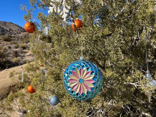 A colorful ornament hangs on a desert tree, surrounded by orange and silver decorations against a clear blue sky.