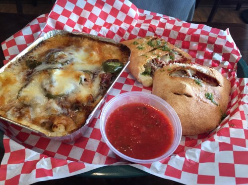 A tray with baked pasta topped with cheese, a side of marinara sauce, and two stuffed bread rolls on a checkered paper.