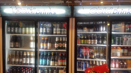 Two glass-fronted refrigerators filled with various cold drinks, including beers and soft drinks, under bright lights.