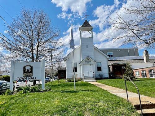 A white town hall building with a bell tower, surrounded by grass and trees, under a blue sky with clouds.