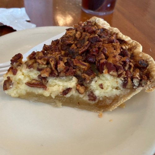 A slice of pecan pie on a plate, featuring a golden crust and a rich filling topped with chopped pecans.