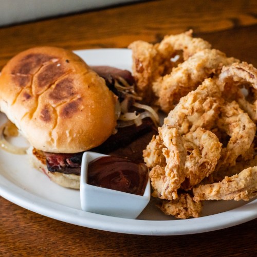 A plate featuring a sandwich with meat and onions, accompanied by crispy onion rings and a small dish of sauce.