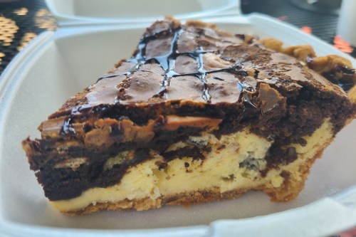 A slice of layered brownie cheesecake with a glossy top, served in a takeout container.
