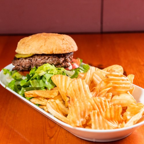 A juicy burger with lettuce, tomato, and pickles, served with a side of crispy wavy potato chips on a wooden table.