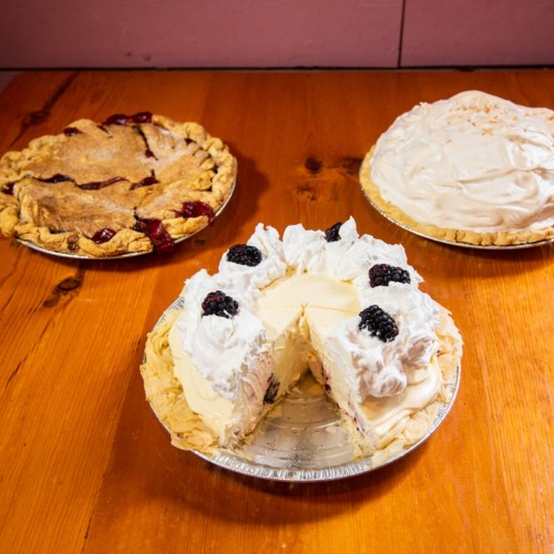 Three pies on a wooden table: a cherry pie, a coconut cream pie, and a sliced cream pie topped with blackberries and whipped cream.