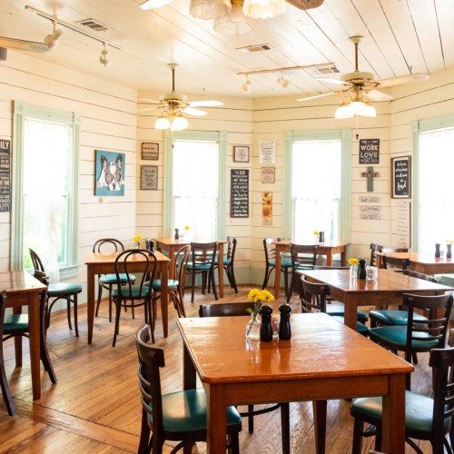 Cozy restaurant interior with wooden tables, chairs, and bright windows, decorated with signs and flowers.