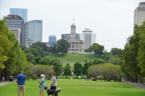 A park scene with people walking a dog, framed by trees, and a historic building on a hill with city skyscrapers in the background.