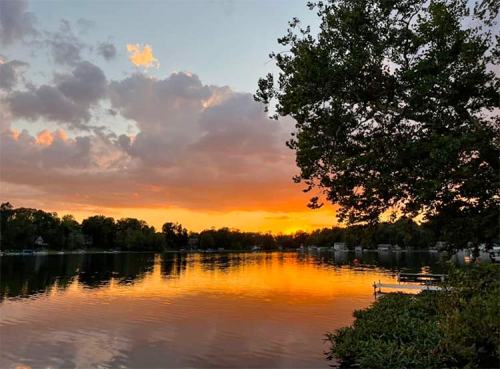 Sunset over a calm lake, with vibrant orange and pink hues reflecting on the water and trees lining the shore.