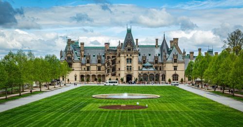 Exterior view of the Biltmore mansion in NC, surrounded by a green lawn and trees, under a cloudy sky.