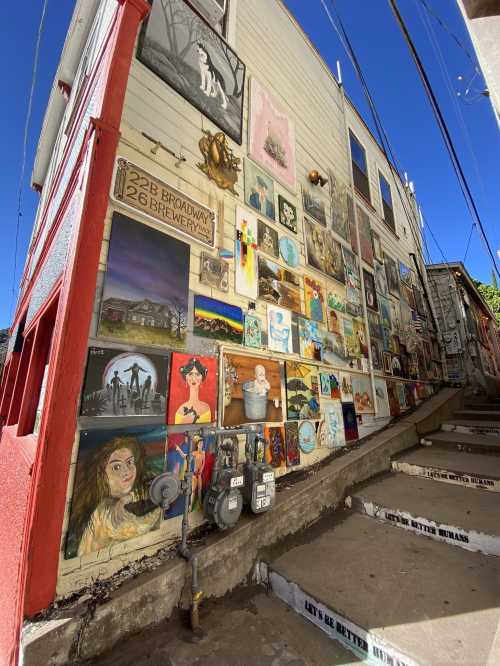 Colorful mural of various artworks displayed on a building wall, with stairs leading down beside it. Clear blue sky above.