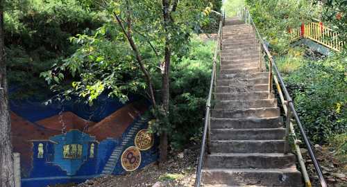 A stone staircase leads up through greenery, with a colorful mural on the left depicting a house and landscape.