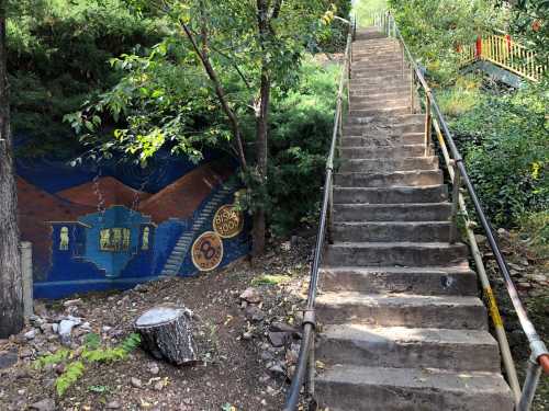 A set of stone stairs leads up through greenery, beside a colorful mural of a house on a hillside.