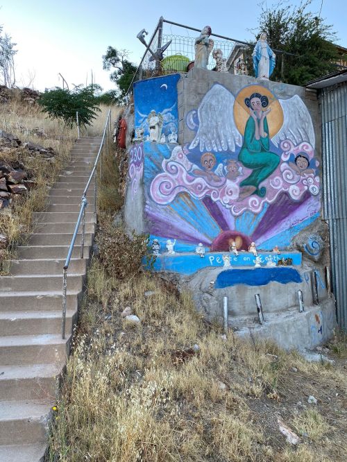 A colorful mural on a hillside, featuring angels and clouds, beside a staircase leading up the slope.