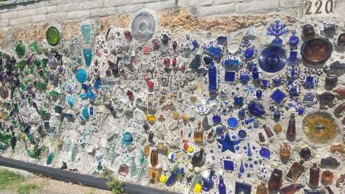 A colorful mosaic wall made of various glass bottles and dishes, featuring blue, green, and clear pieces.