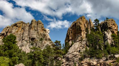 Mount Rushmore with carved faces, surrounded by rocky cliffs and trees under a partly cloudy sky.