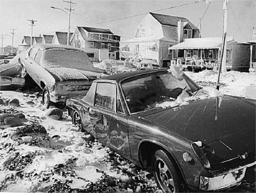 Two snow-covered cars parked on a snowy street, with houses in the background and power lines overhead.