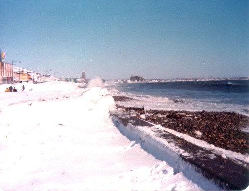 A snowy beach scene with waves crashing, featuring a boardwalk and distant buildings under a clear blue sky.