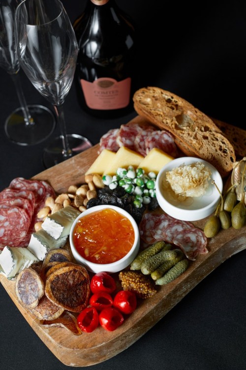 A wooden platter filled with assorted meats, cheeses, pickles, nuts, and spreads, accompanied by wine glasses and a bottle.