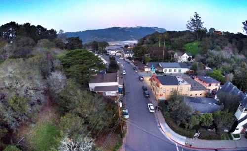 Aerial view of a small town with trees, buildings, and a winding road, set against a backdrop of hills and a clear sky.