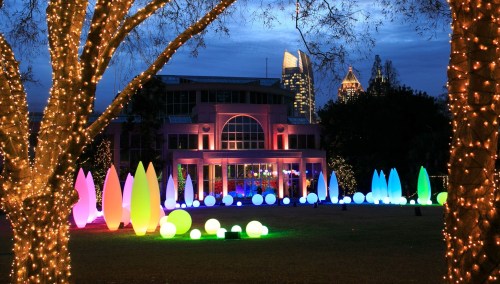 Colorful illuminated sculptures and trees surround a building, with a twilight sky and city skyline in the background.