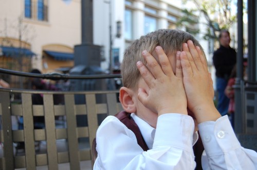 A young boy in a white shirt and vest covers his face with his hands, sitting at an outdoor café.