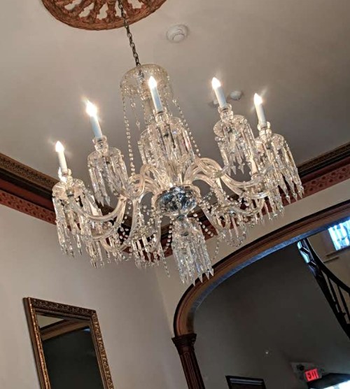 A sparkling crystal chandelier with six candle-like lights hangs from a decorative ceiling in a well-lit hallway.
