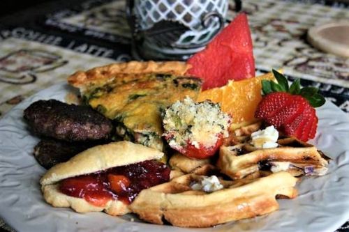 A plate of breakfast featuring waffles, sausage, a vegetable quiche, fruit slices, and a pastry with jam.